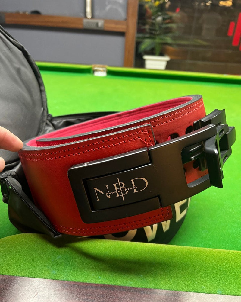 Red leather NBD lever belt being packed into the NBD round belt bag on a green table, with POWERLIFTING lettering on the back of the bag