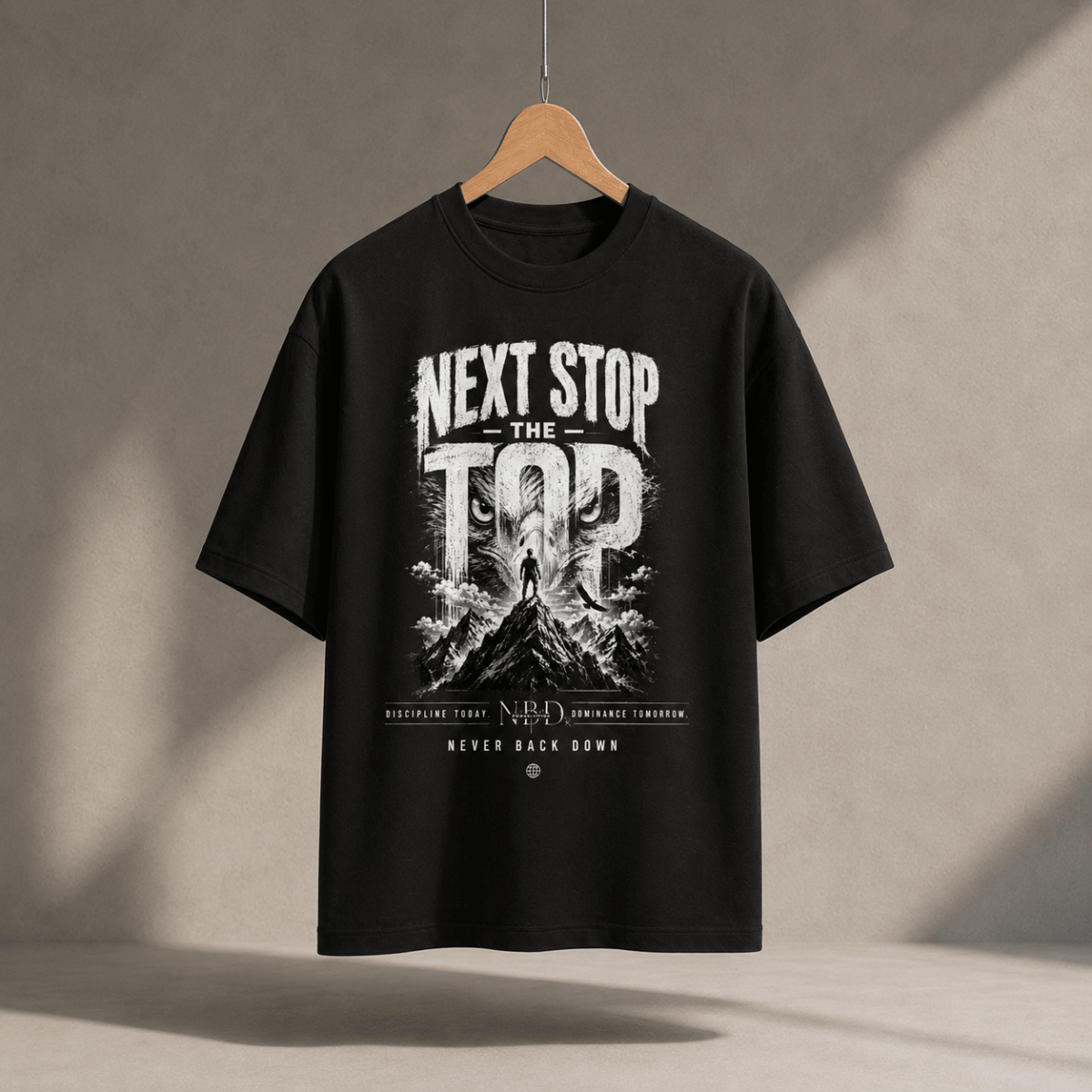 NBD 'Next Stop The Top' oversized T-shirt in black, hanging on a wooden hanger, front print of a lifter on a mountain summit with 'Never Back Down' below